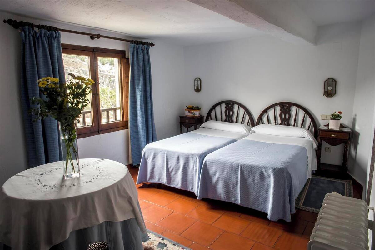 A room with two beds, a round table with a vase, and a window with curtains.