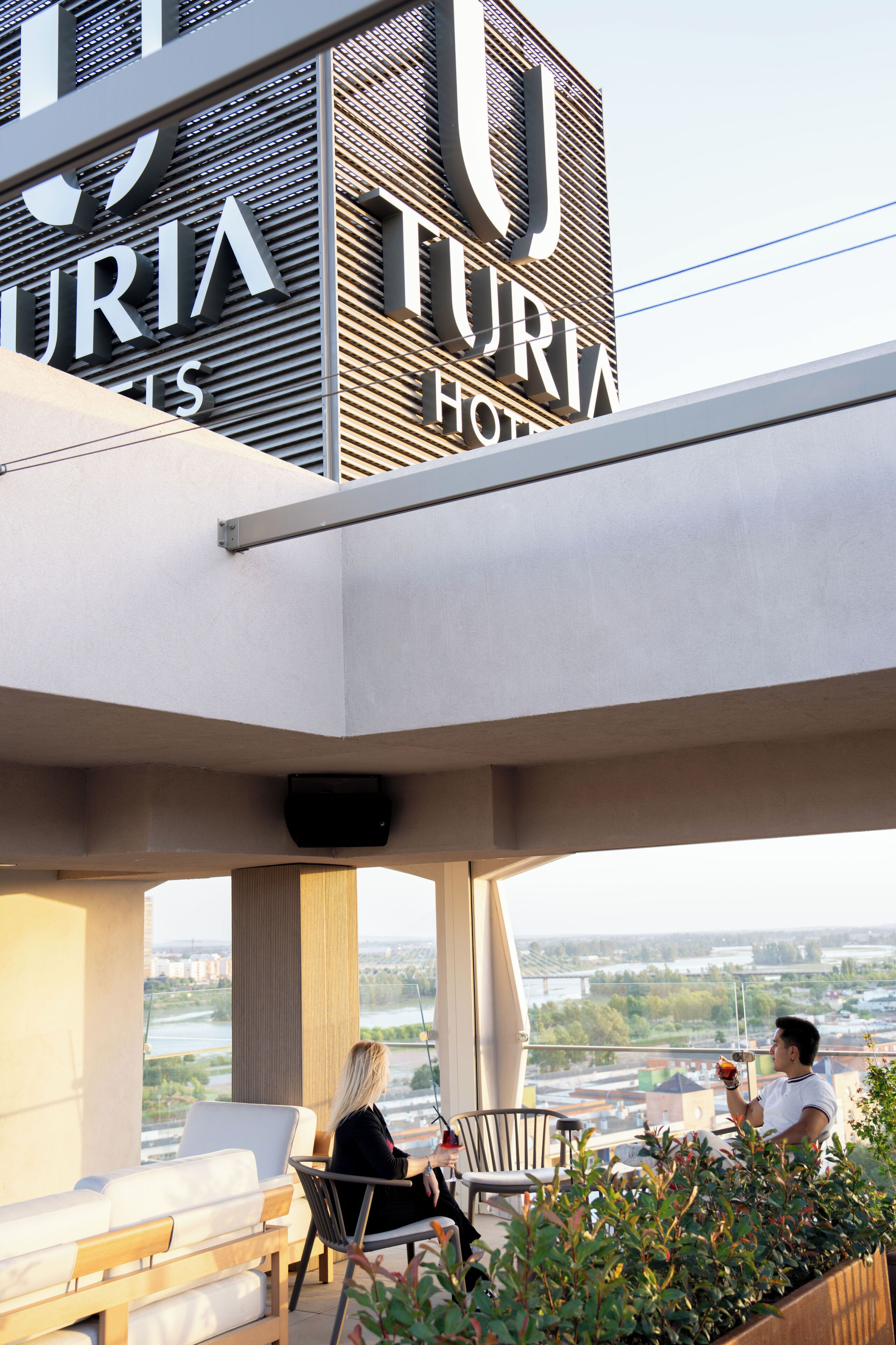 Un hotel moderno con un gran letrero que dice 'TURIA HOTEL', zona de estar al aire libre con vistas a la ciudad y un área de balcón con plantas.