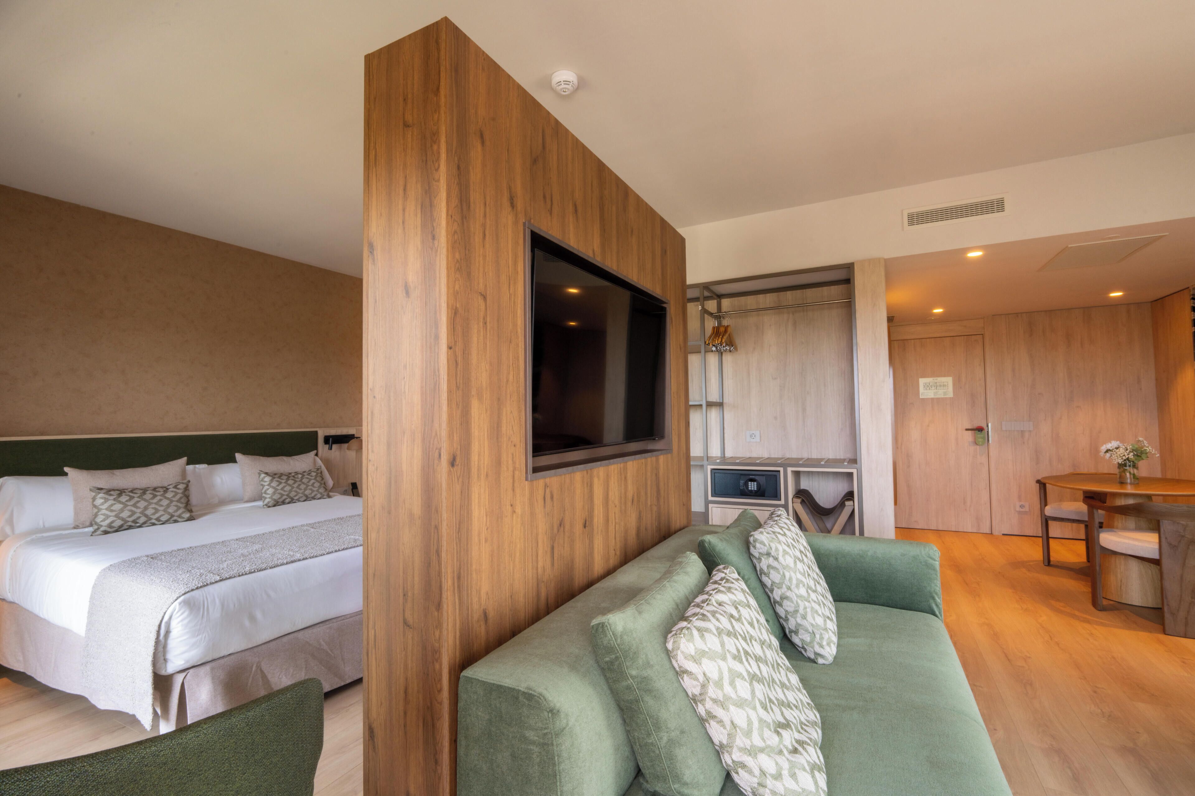A hotel room with a bed, a green sofa, a TV mounted on the wall, and a kitchenette area.