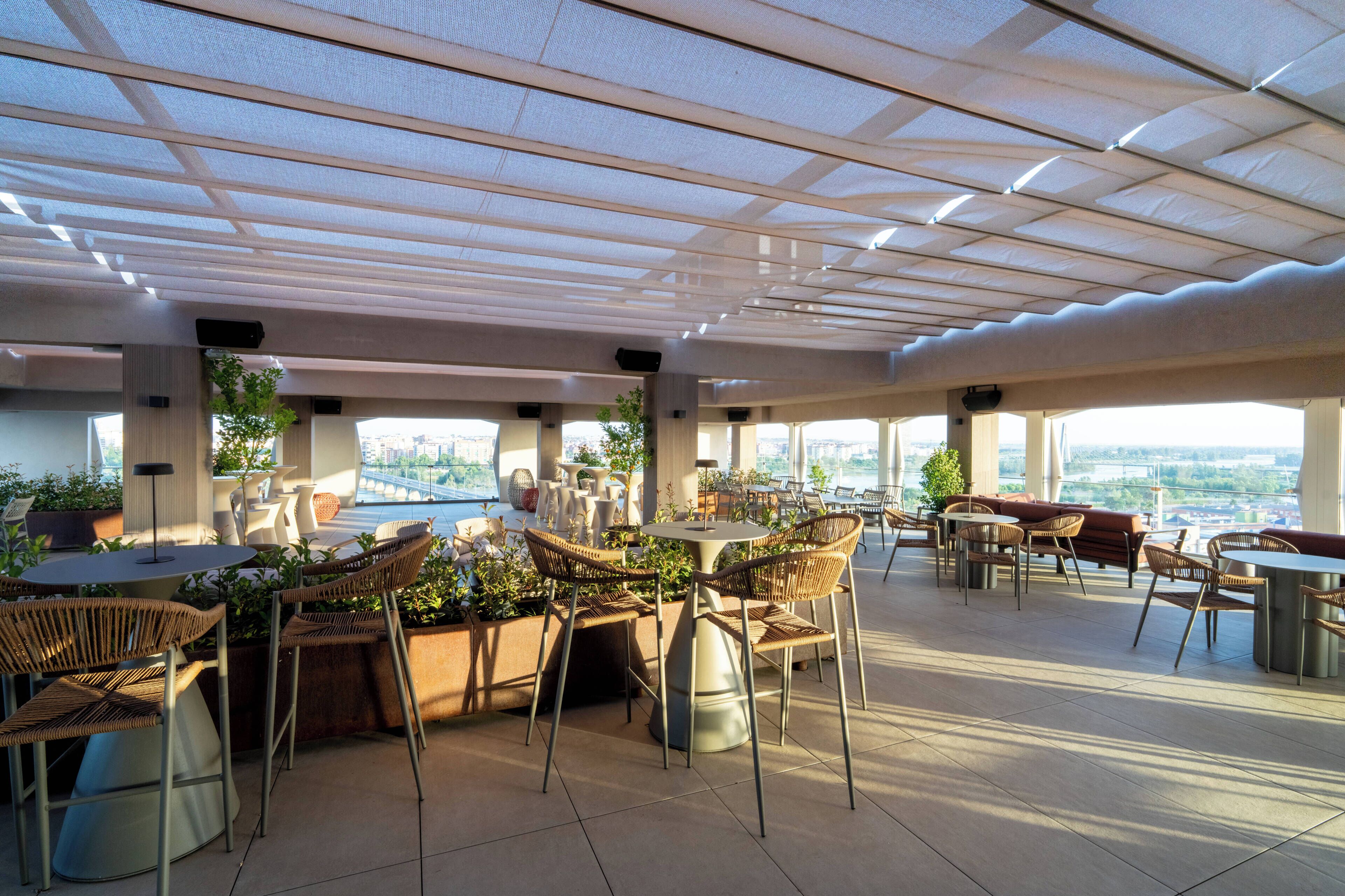 A rooftop terrace with outdoor seating, potted plants, and large windows offering city views.