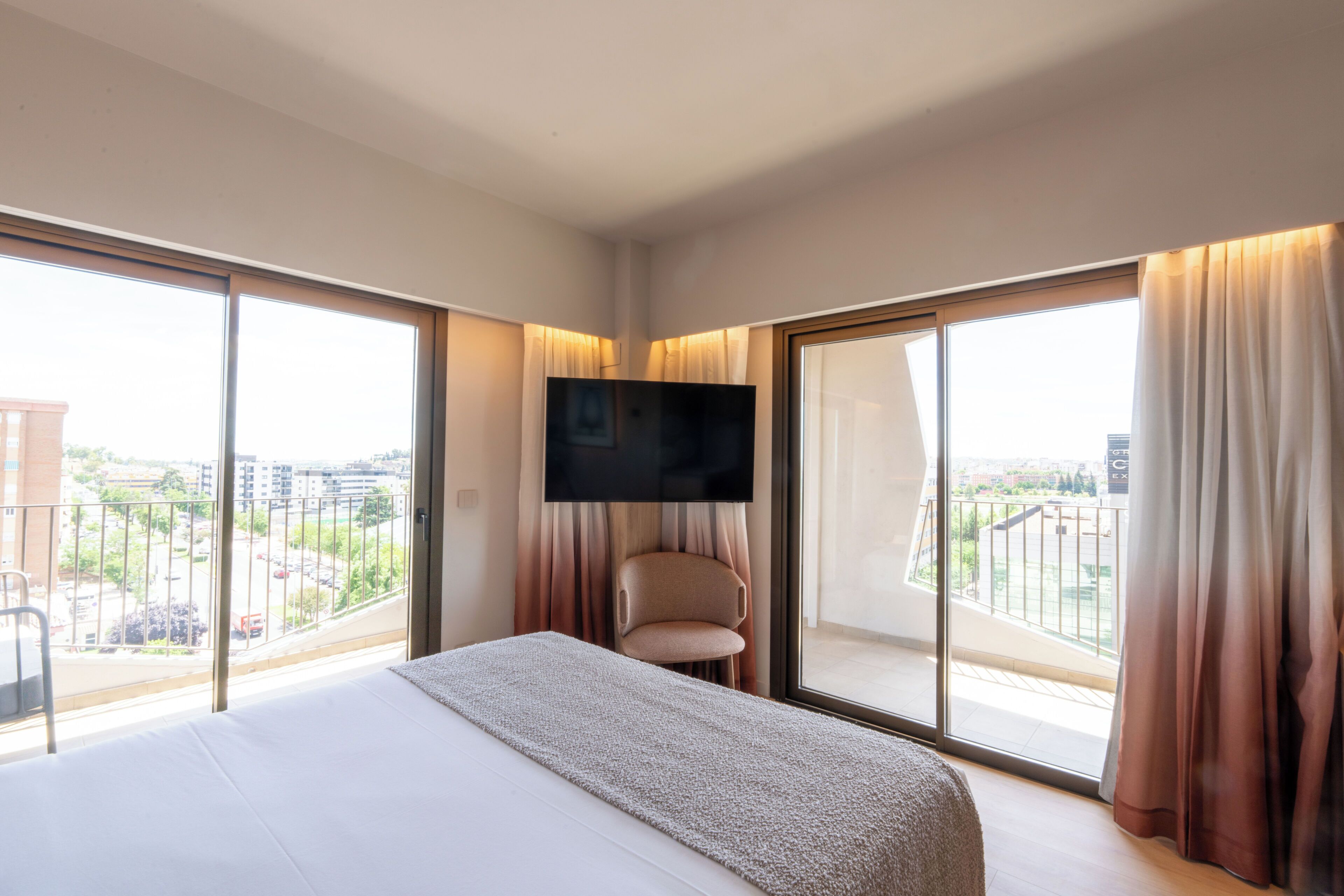 Habitación de hotel con una cama grande, un televisor de pantalla plana en la pared, una silla y un balcón con cortinas.