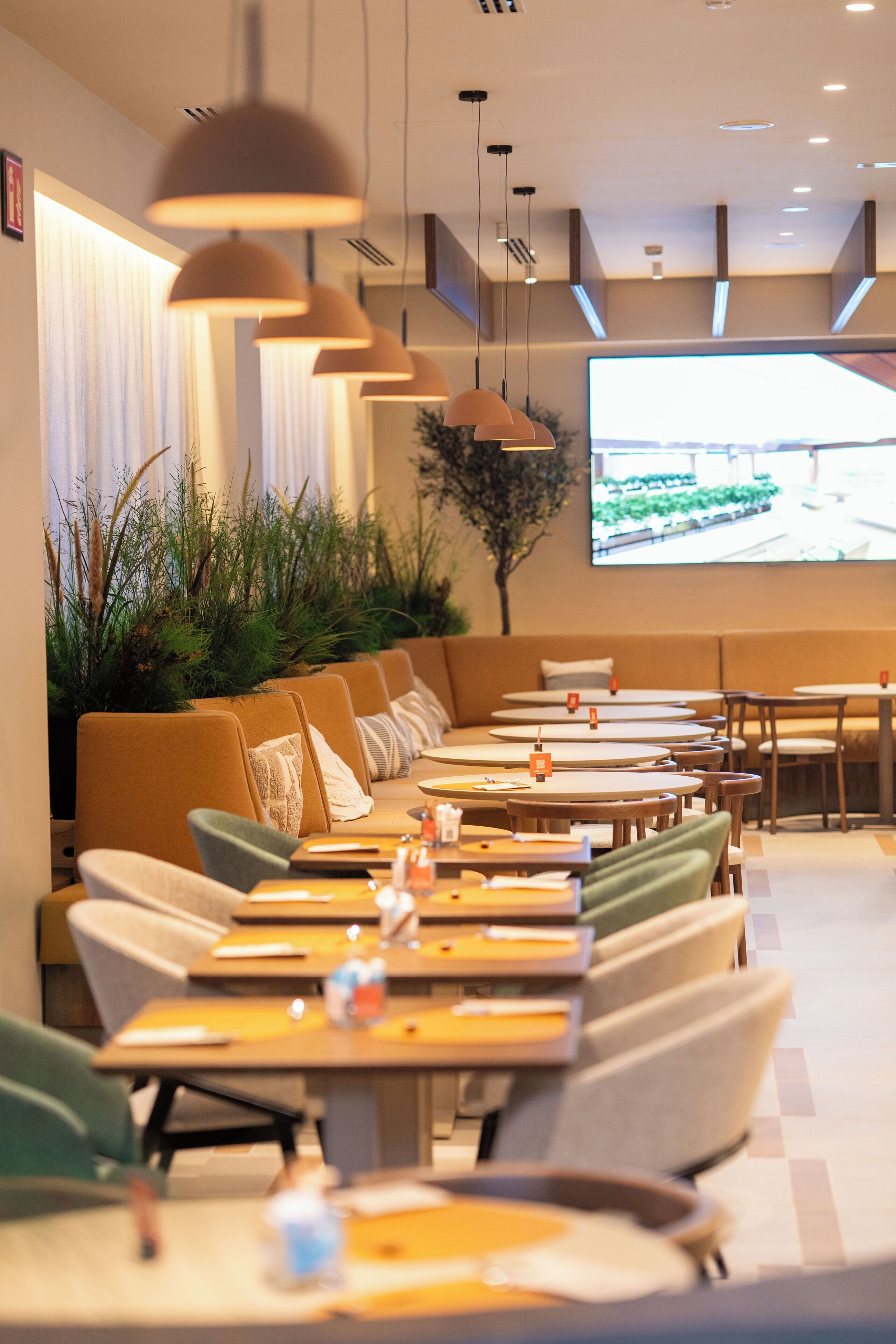 A modern restaurant with a long counter, cushioned seating, pendant lighting, and a large screen displaying an outdoor scene.