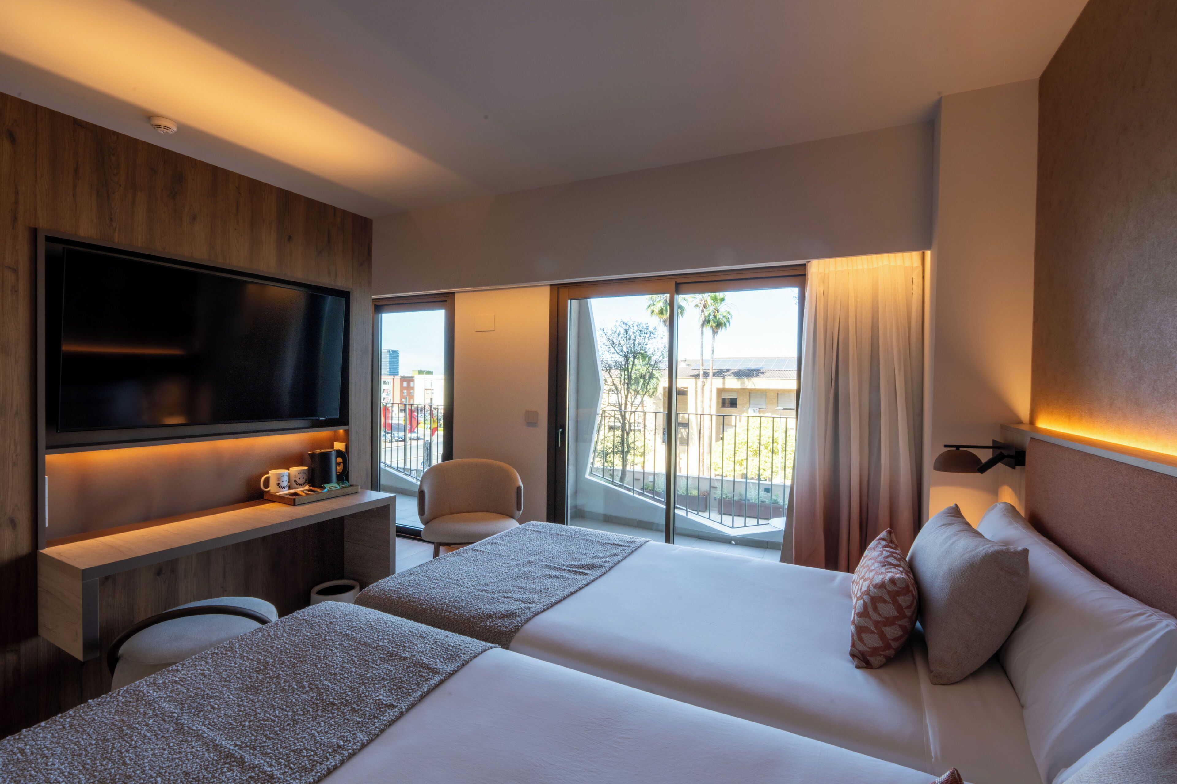 A hotel room with a large flat-screen TV, a sofa, a bed with two pillows, a small table with a cup, and a view of the outside.