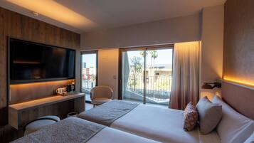 A hotel room with a large flat-screen TV, a sofa, a bed with two pillows, a small table with a cup, and a view of the outside.