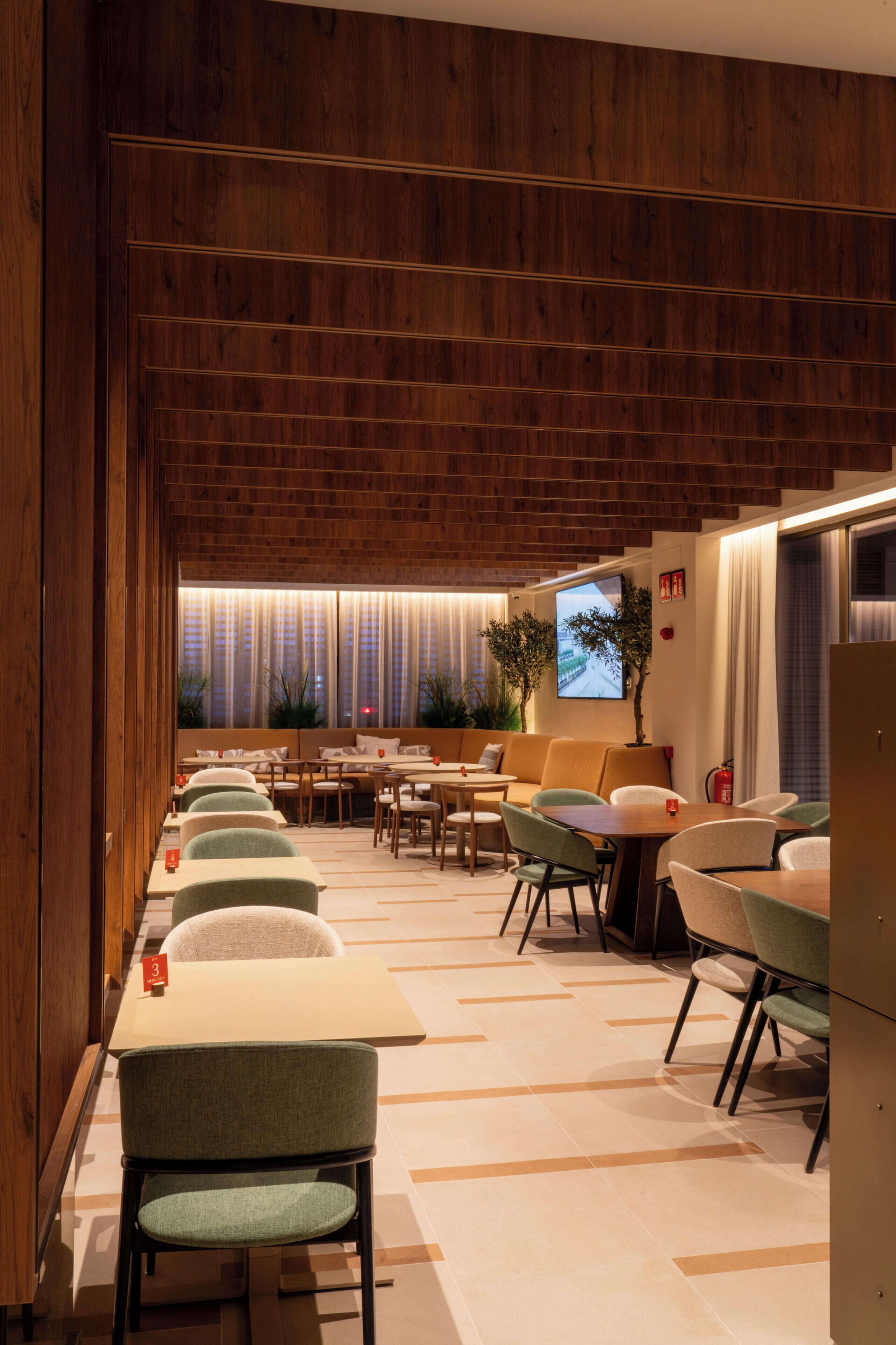 A modern hotel lobby with wooden paneling, a seating area with tables and chairs, a television screen, and a plant.