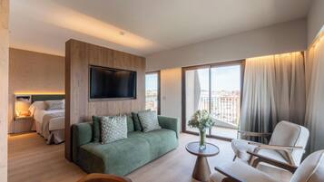 Una habitación de hotel moderna con un sofá verde, una mesa de centro de madera, un televisor montado en la pared, una cama con una mesita de noche y vistas a la ciudad a través de amplias ventanas.