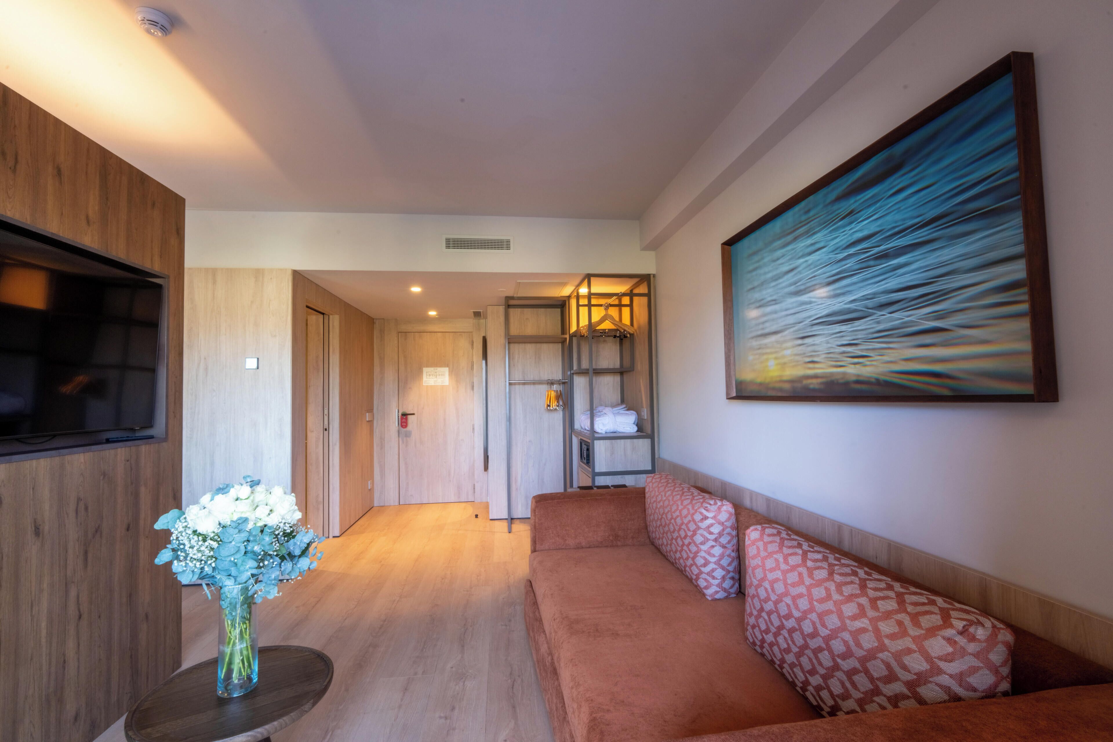 A hotel room with a wooden floor, a sofa with patterned cushions, a flat-screen TV, a vase with flowers, and a painting on the wall.