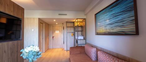 A hotel room with a wooden floor, a sofa with patterned cushions, a flat-screen TV, a vase with flowers, and a painting on the wall.