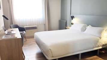 Habitación de hotel con una cama grande, un escritorio con una computadora portátil, una silla, un radiador y una ventana con cortinas.