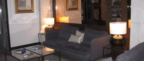 A living room with a sofa, a coffee table, and two lamps. There are framed artworks on the wall and a mirror reflecting the room.