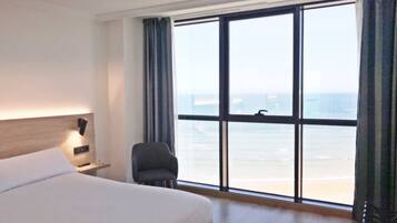 Una habitación de hotel con un ventanal grande, una cama, un sillón y una mesita de noche.