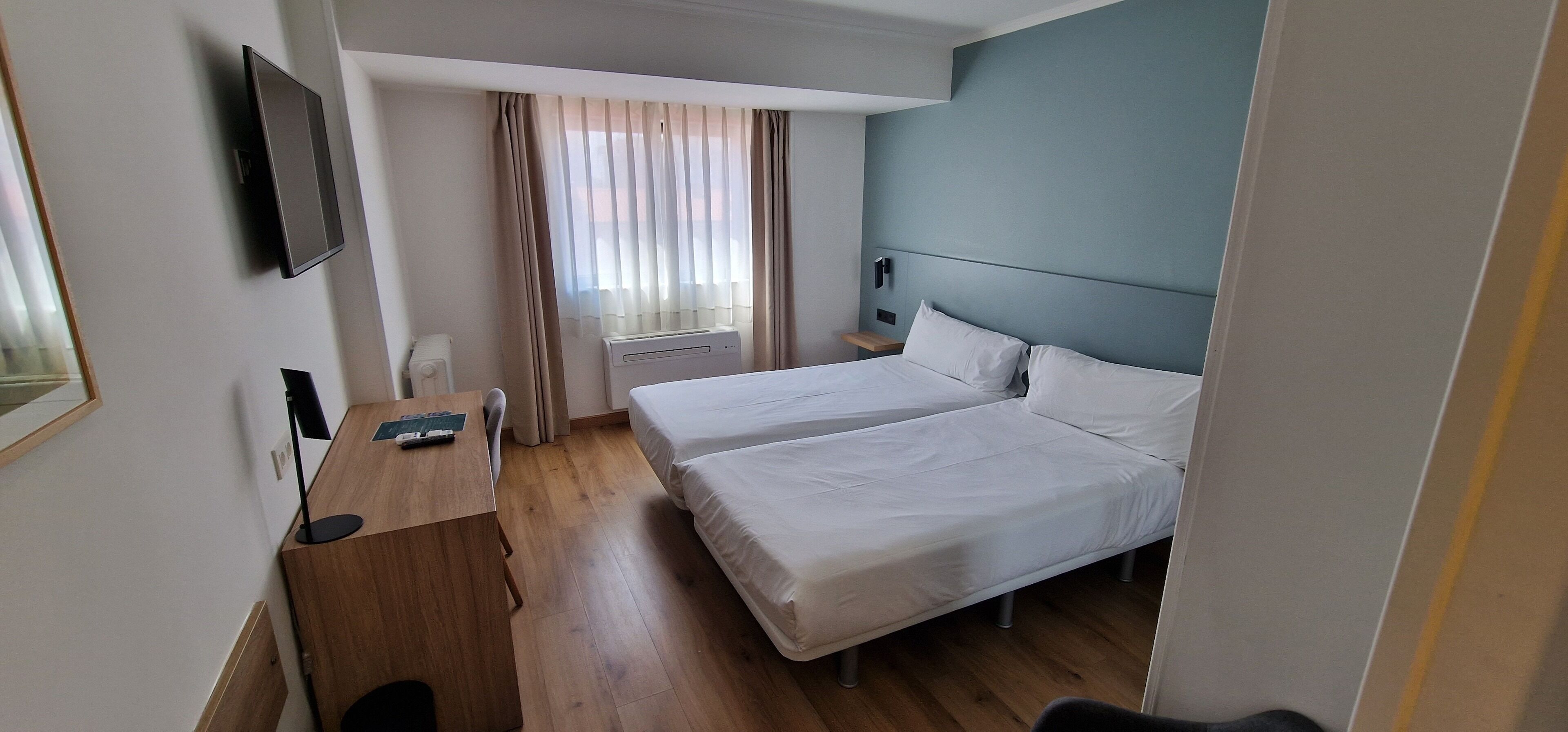 A hotel room with two beds, a desk with a chair, a TV mounted on the wall, and a window with curtains.