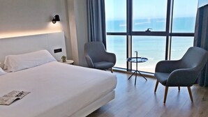 Ein Hotelzimmer mit einem großen Bett, zwei Sesseln, einem kleinen Tisch und Meerblick durch große Fenster.