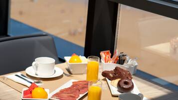 A breakfast table with a view of the beach, including a plate of food, a cup, a glass of orange juice, and a bowl of fruit.