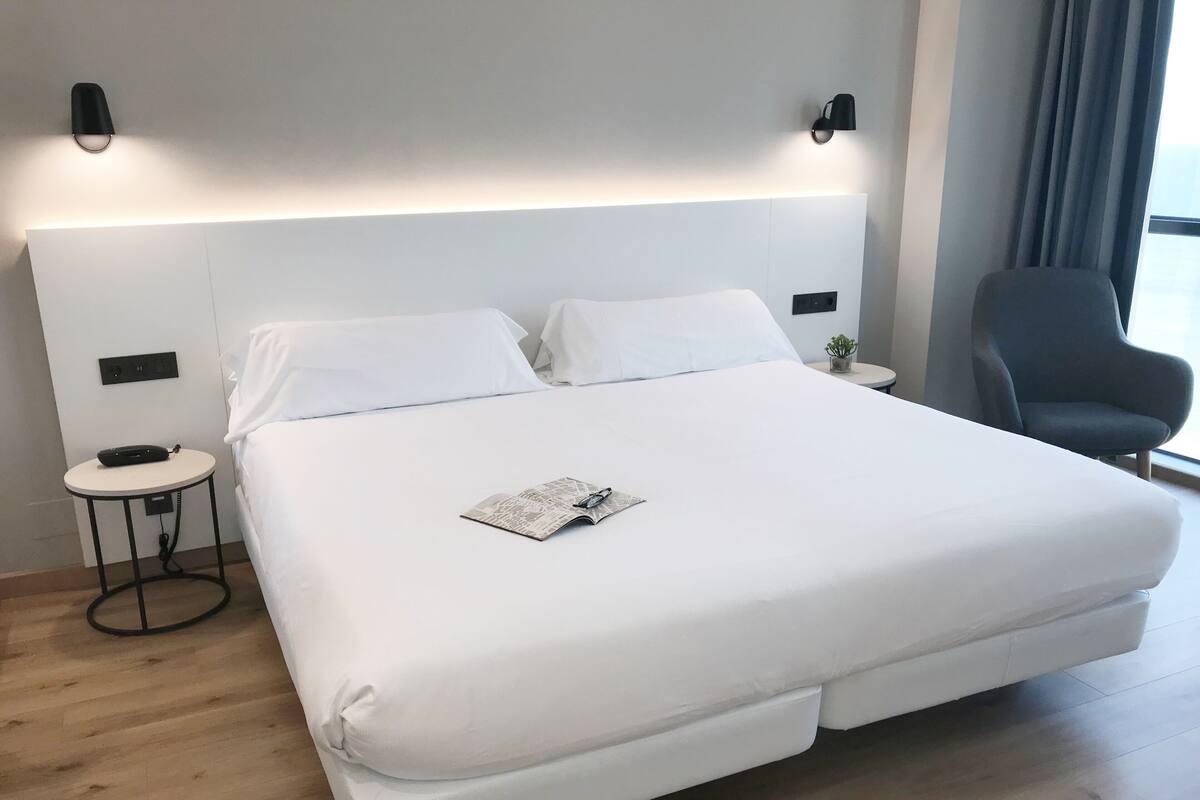 Una habitación de hotel moderna con una cama grande, mesitas de noche, una silla y un ventanal con cortinas.