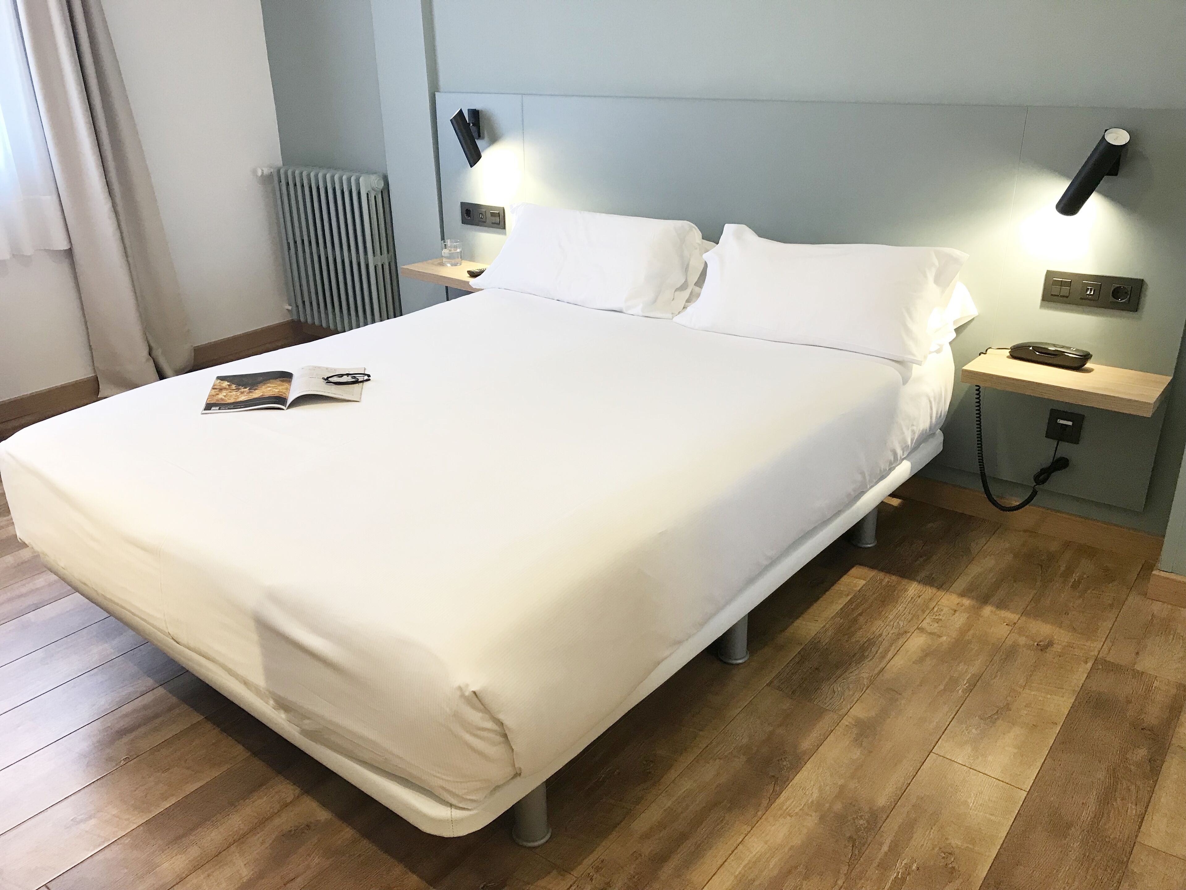 Un lit bien fait, avec du linge de lit blanc, une table de chevet avec une lampe, un téléphone et un magazine posés sur le lit.