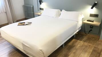 A neatly made bed with white linens, a bedside table with a lamp, a phone, and a magazine on the bed.