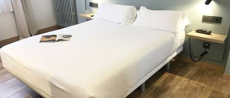 Un lit bien fait, avec du linge de lit blanc, une table de chevet avec une lampe, un téléphone et un magazine posés sur le lit.