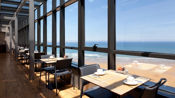 Ein Restaurant mit Blick auf den Strand. Tische stehen entlang der Fenster, mit Stühlen und Esstischen bestückt.