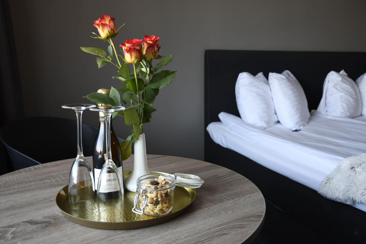 Habitación de hotel con cama, almohadas, un jarrón con rosas, copas de vino y un recipiente con nueces sobre una bandeja.