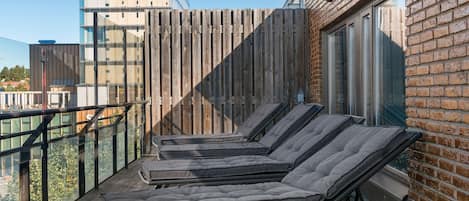A balcony with four sun loungers, a wooden fence, and a brick wall with a window.