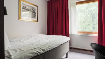 A single bed with a white duvet, a framed photograph on the wall, a window with red curtains, and a black chair.