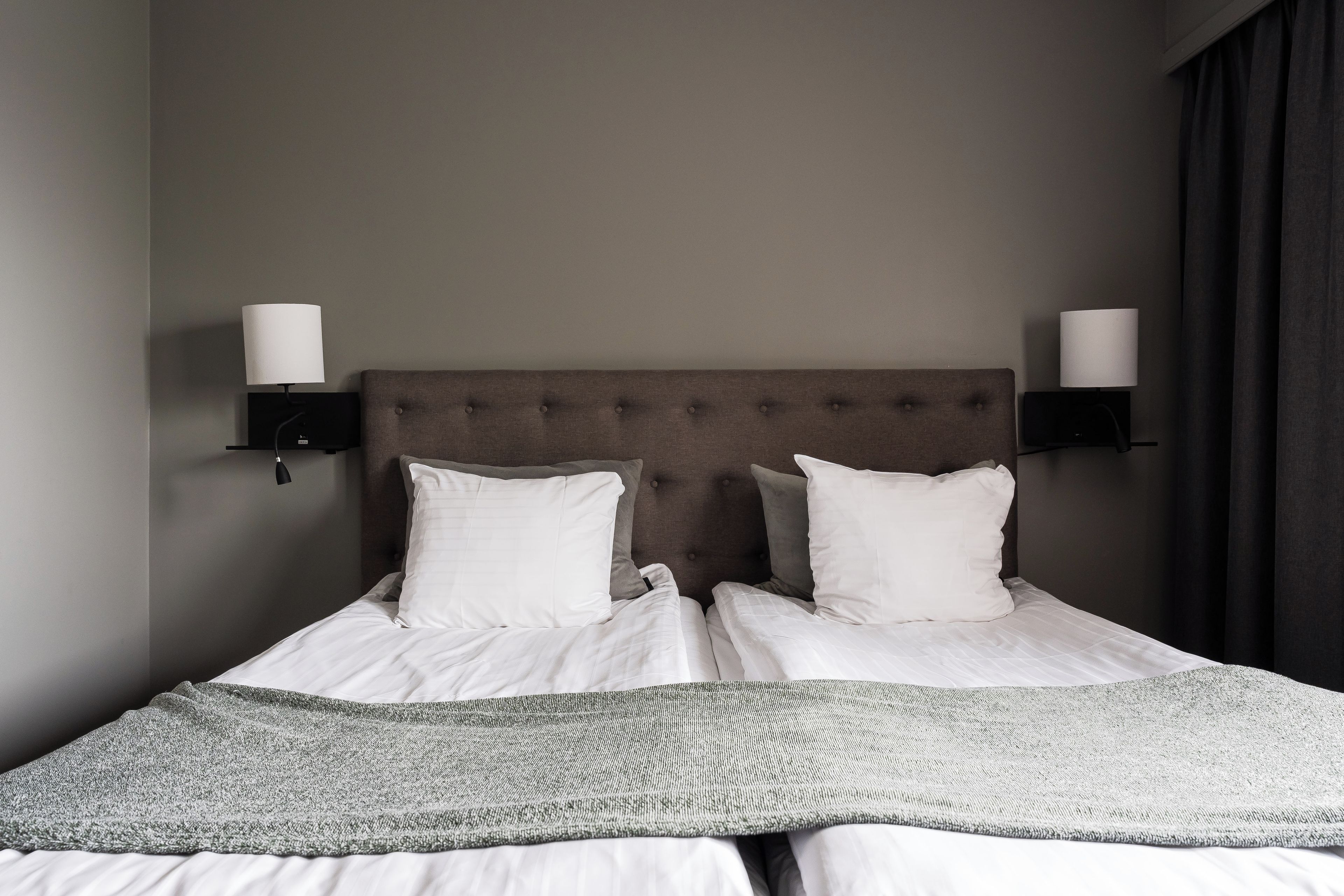 Una cama bien tendida con sábanas blancas y un plaid de punto gris, flanqueada por dos lámparas fijadas a la pared y una cortina oscura.