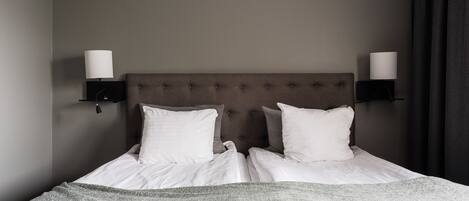 Una cama bien tendida con sábanas blancas y un plaid de punto gris, flanqueada por dos lámparas fijadas a la pared y una cortina oscura.