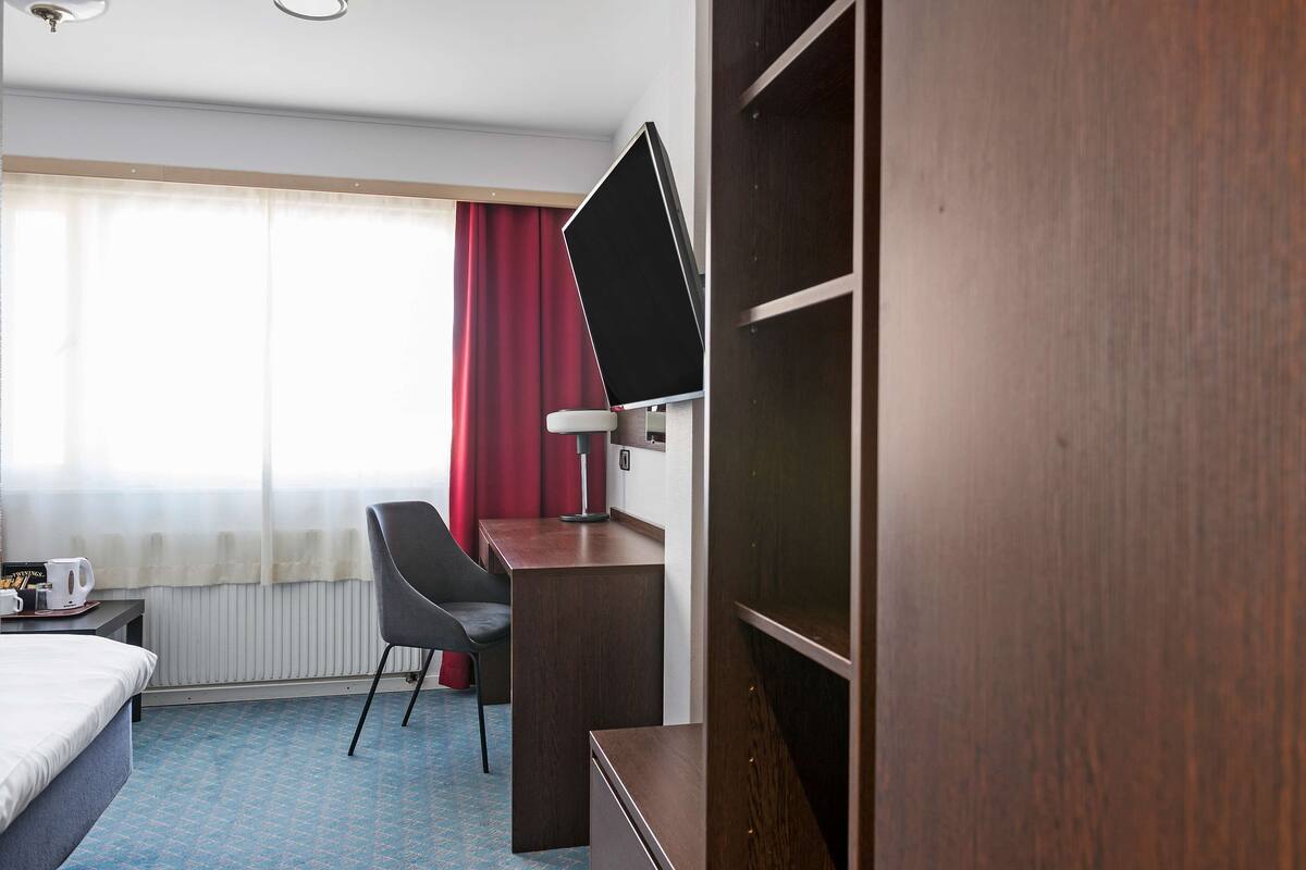 Habitación de hotel con cama, escritorio, silla, televisión de pantalla plana, radiador y ventana con cortinas.