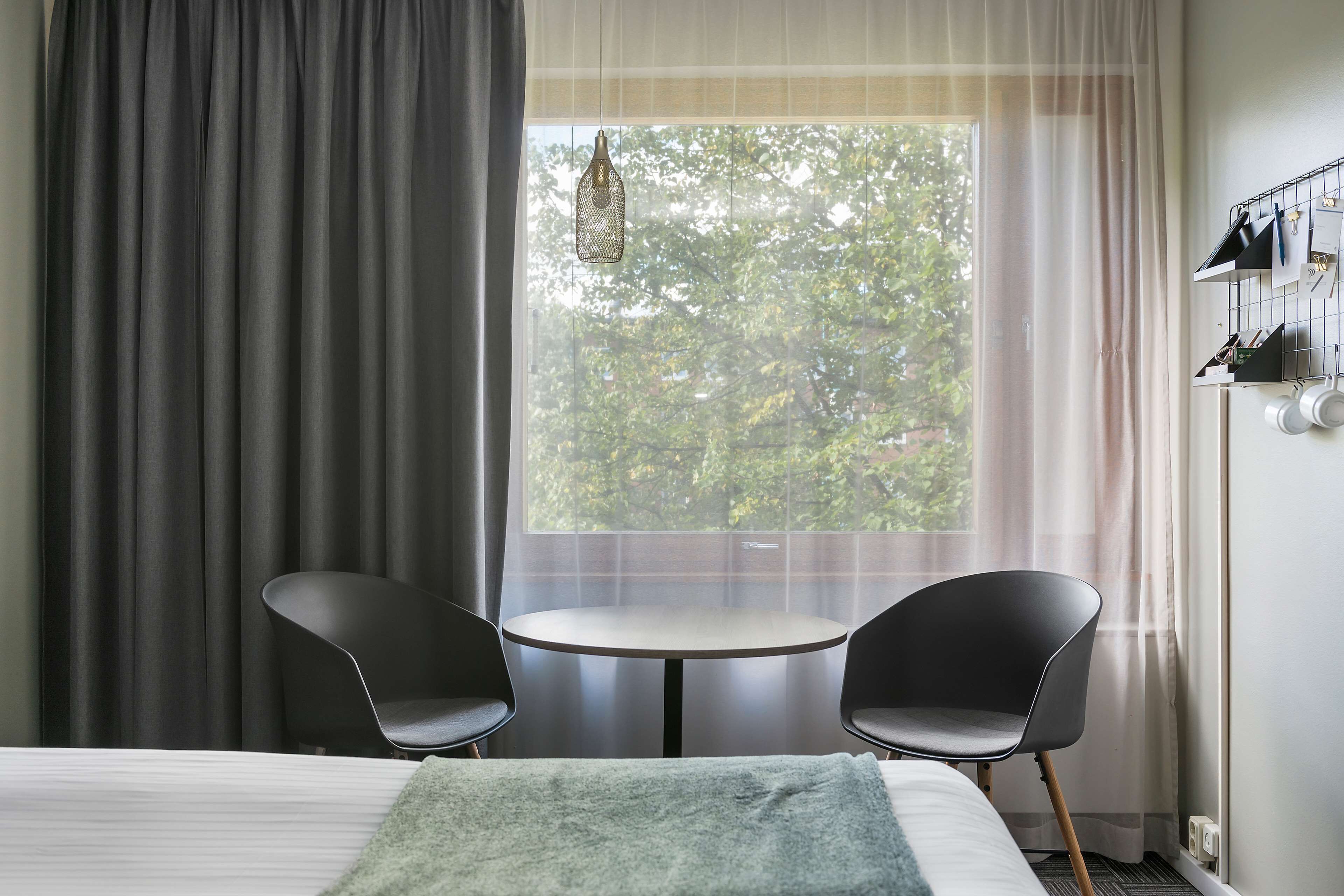 Habitación de hotel con cama, dos sillas negras, una mesita redonda, ventana con cortinas y una lámpara colgante.