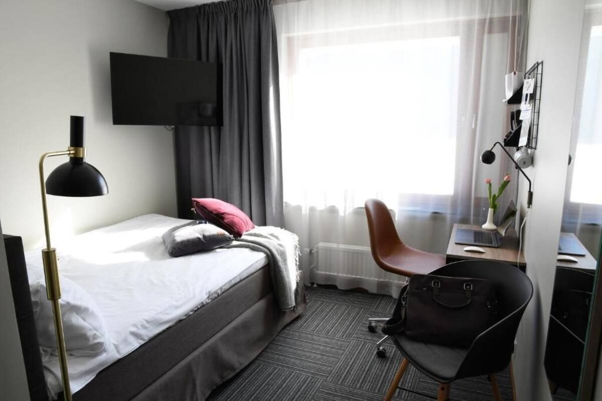 Una habitación de hotel con una cama, un escritorio con silla, un televisor en la pared y una ventana con cortinas.