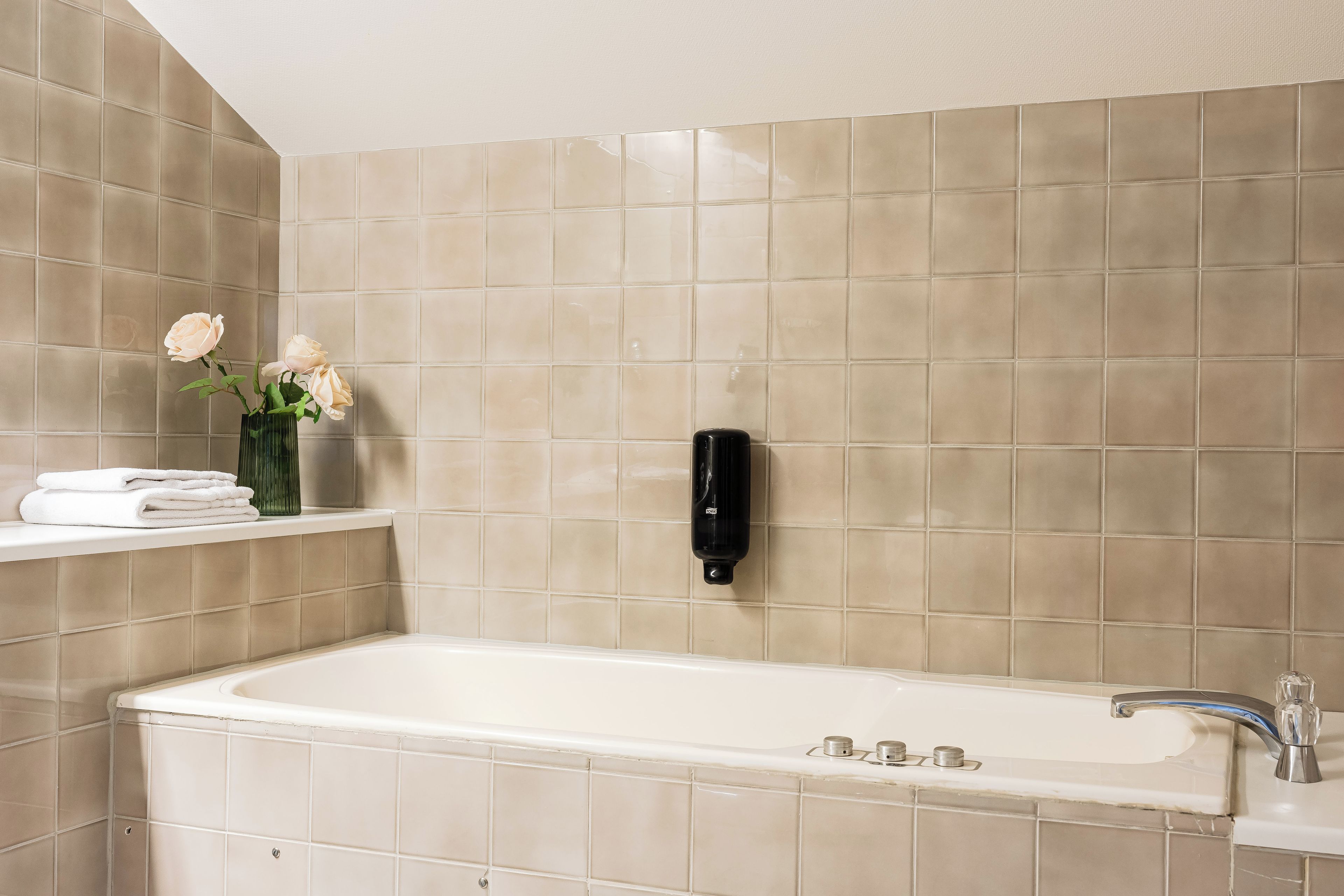 A bathroom with a white bathtub, beige tiles, a towel rack with folded towels, a vase with flowers, and a black soap dispenser.