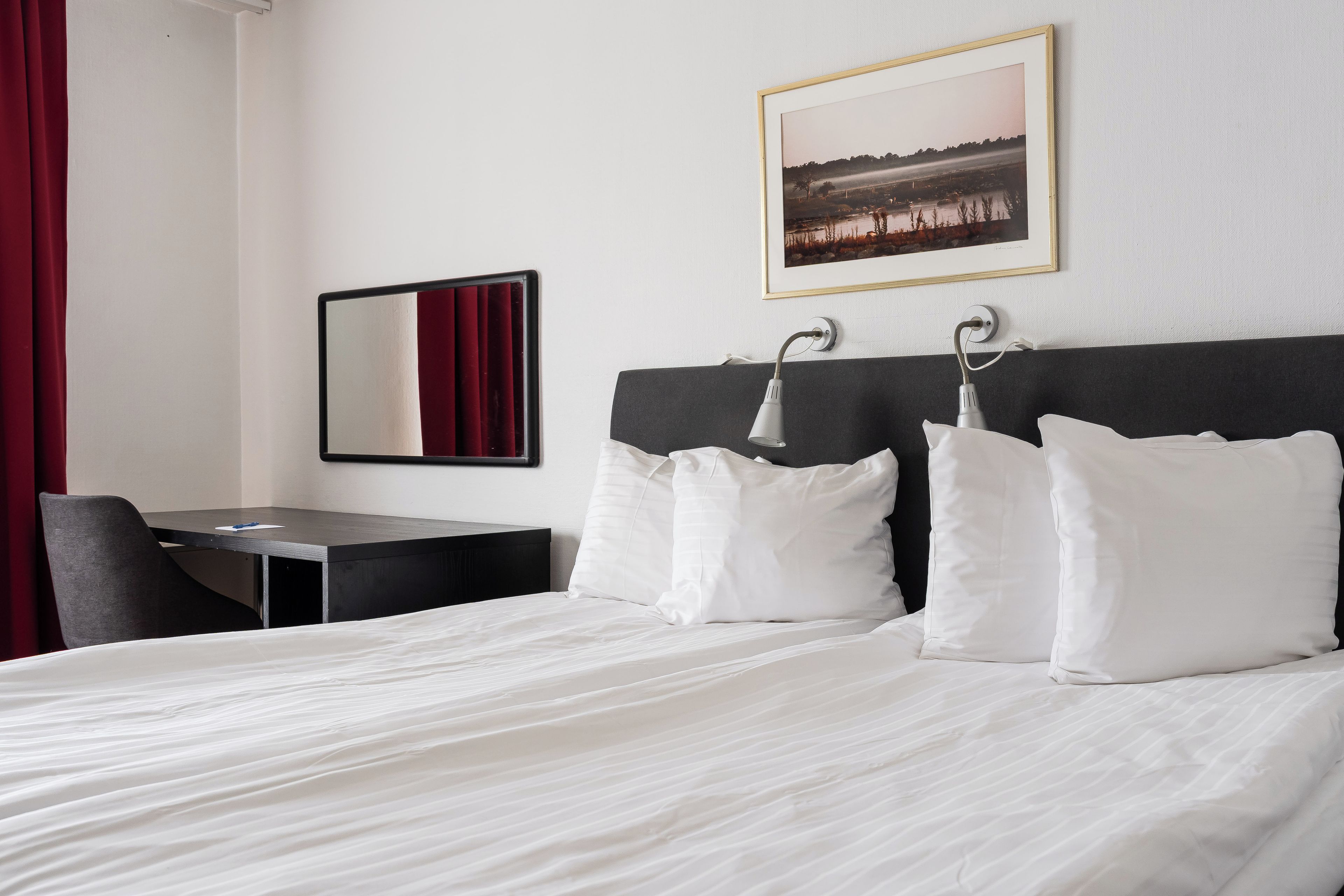 A hotel room with a bed, a desk, a chair, a mirror, and a framed picture on the wall.