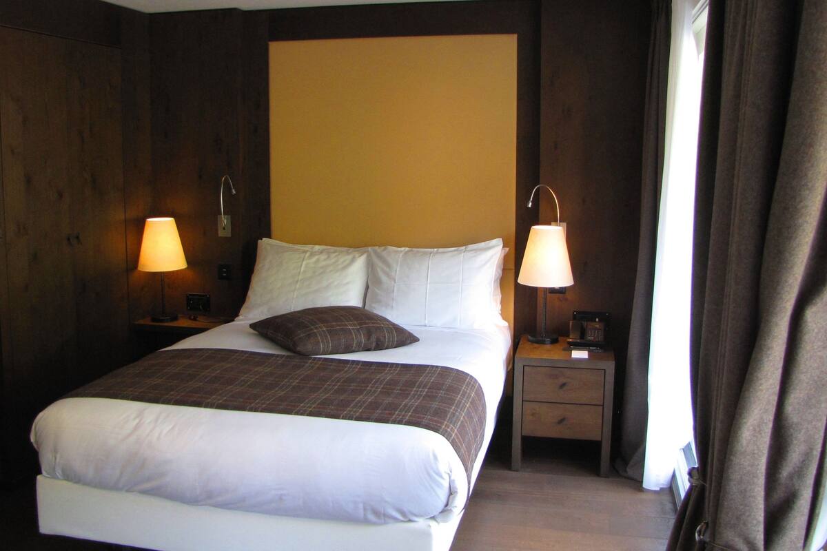 Una cama bien hecha con sábanas blancas y una manta marrón de cuadros, flanqueada por dos mesitas de noche con lámparas, en una habitación con paneles de madera y un gran ventanal con cortinas.