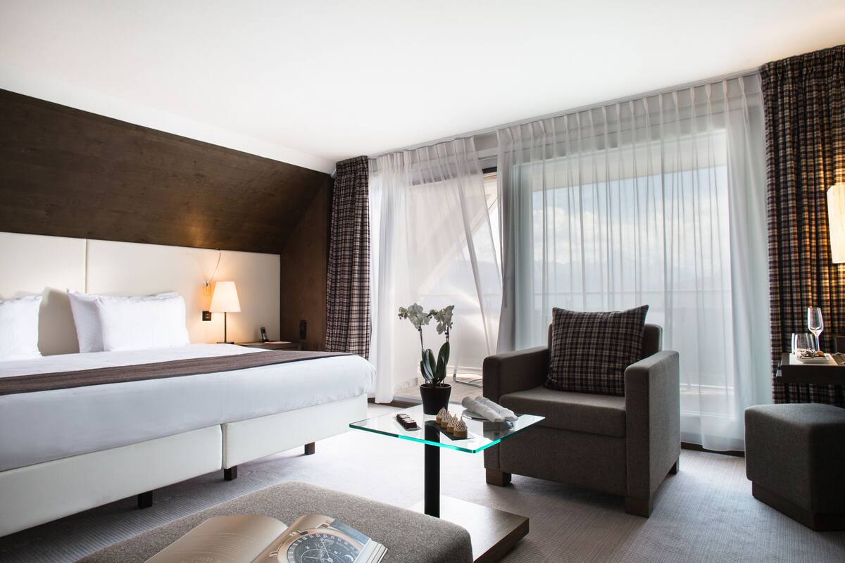Una habitación de hotel con una cama, un sofá, una mesita de vidrio, una pequeña mesa con una lámpara y una planta en maceta.
