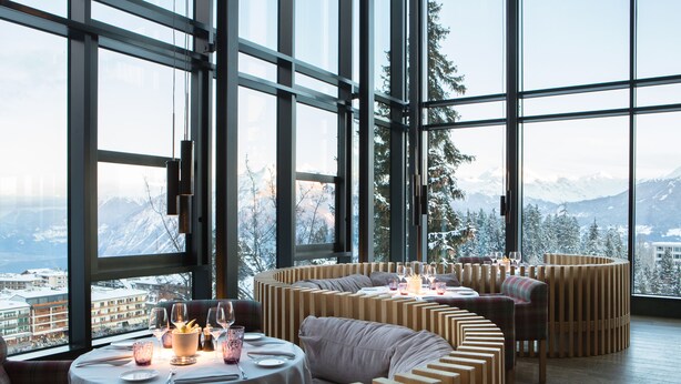 Un interior moderno con ventanas amplias, mobiliario de madera y vistas a montañas nevadas.