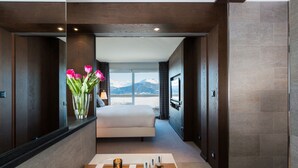 Una habitación de hotel moderna con una cama grande, un tocador de madera con artículos de aseo, vistas a montañas y un espejo amplio.