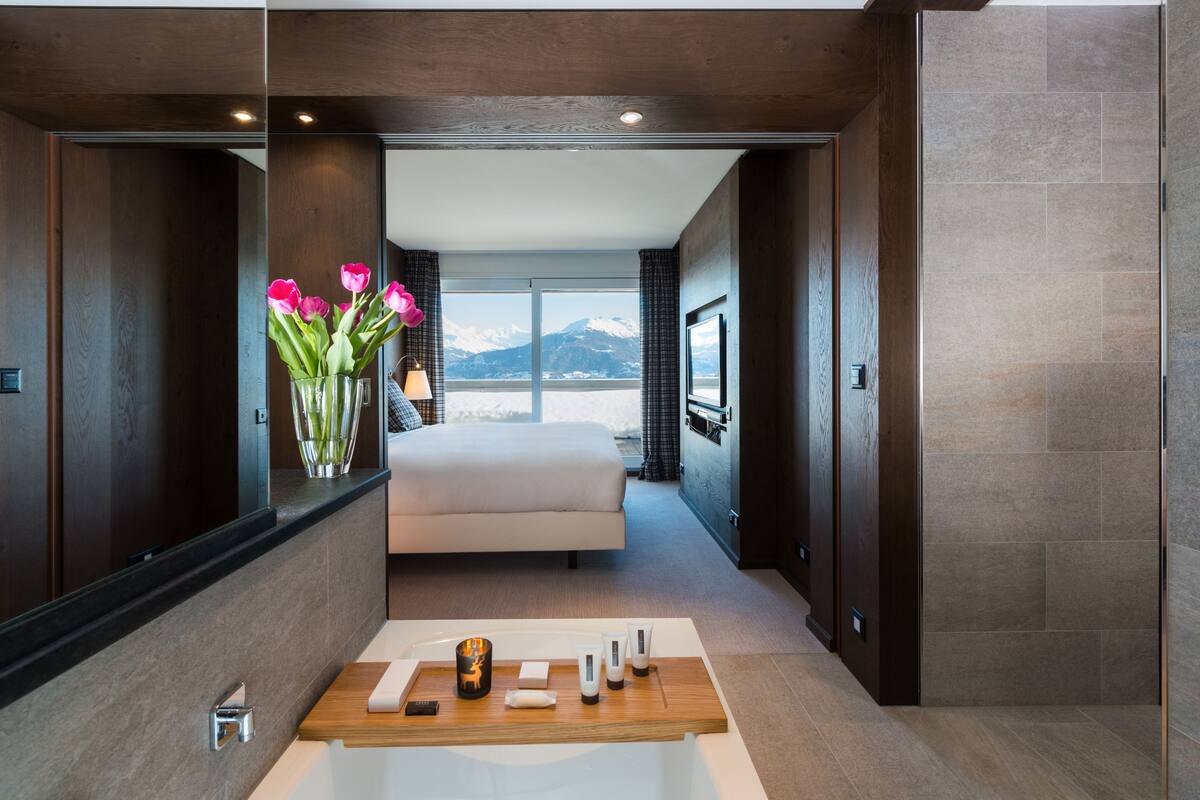 Una habitación de hotel moderna con una cama grande, un tocador de madera con artículos de aseo, vistas a montañas y un espejo amplio.