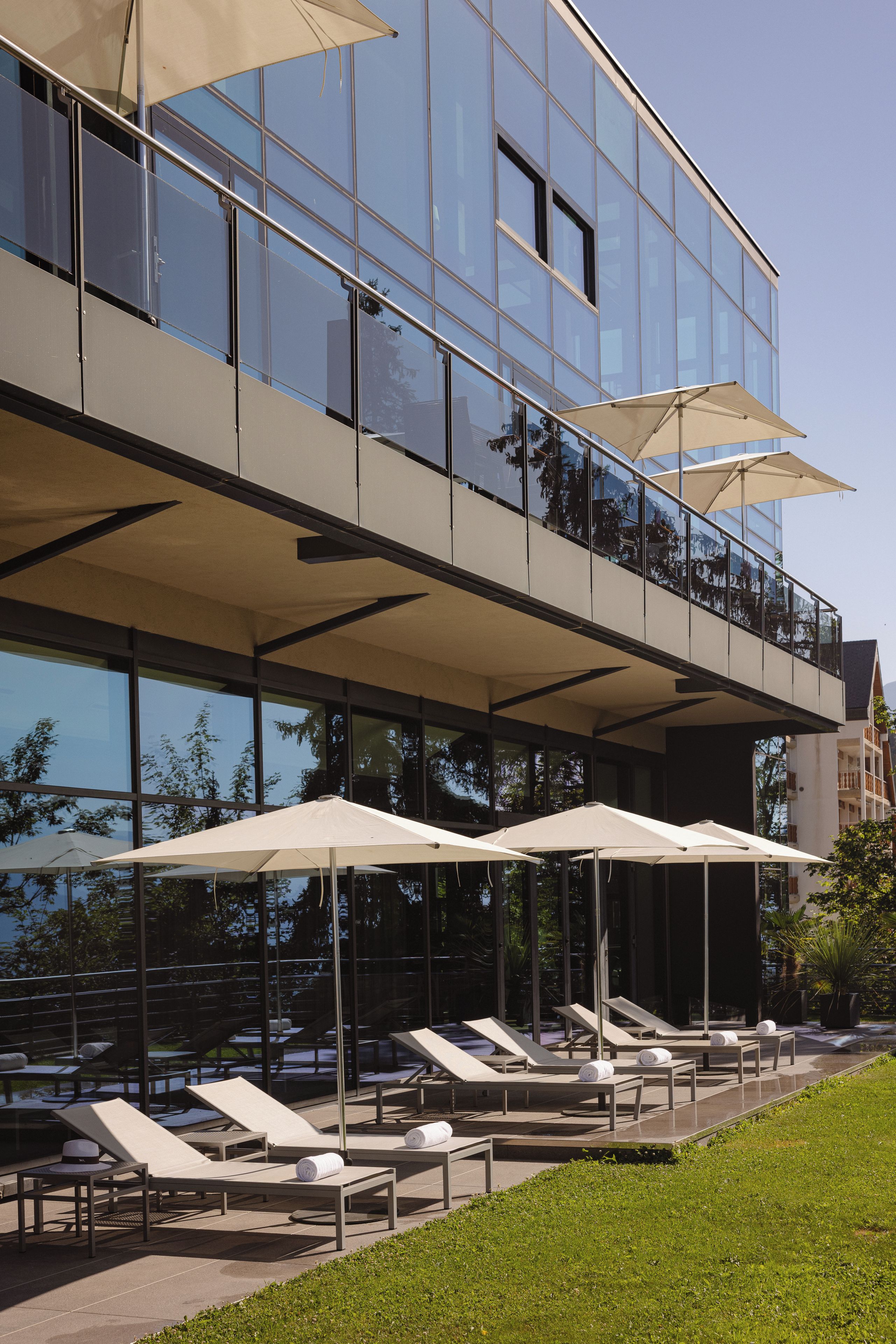 A modern building with a glass facade, outdoor seating with umbrellas, and a pool area.
