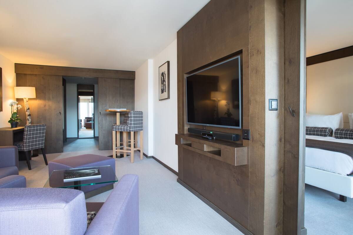 Una habitación de hotel moderna con una televisión de pantalla plana montada en un panel de madera, un sofá, una mesa de centro de vidrio y una cama con ropa de cama escocesa.
