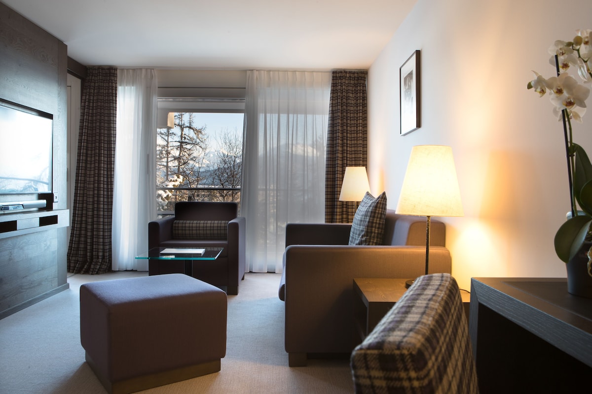 Habitación de hotel con sofá, reposapiés y una mesa con lámpara. Se ve una ventana con cortinas y vistas a árboles.