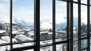 Vista a través de amplias ventanas de un pueblo cubierto de nieve con montañas al fondo.