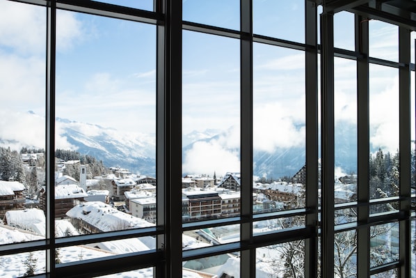 Vista a través de amplias ventanas de un pueblo cubierto de nieve con montañas al fondo.