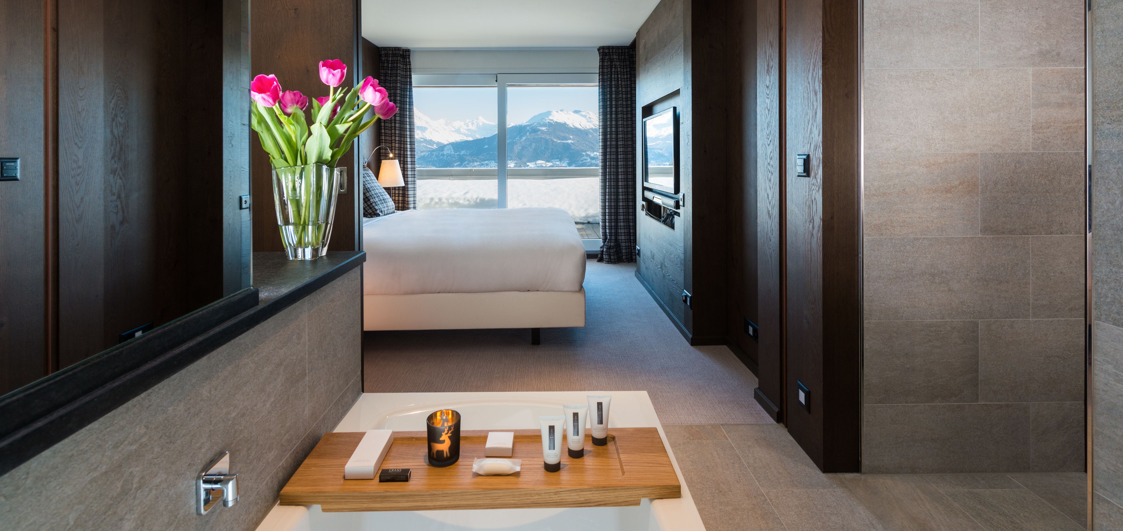 Une chambre d’hôtel moderne avec un grand lit, une vue sur les montagnes, une table de chevet en bois avec des articles de toilette et un vase de fleurs.