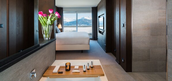 Una habitación de hotel moderna con una cama grande, vistas a las montañas, una mesita de madera con artículos de aseo y un jarrón con flores.
