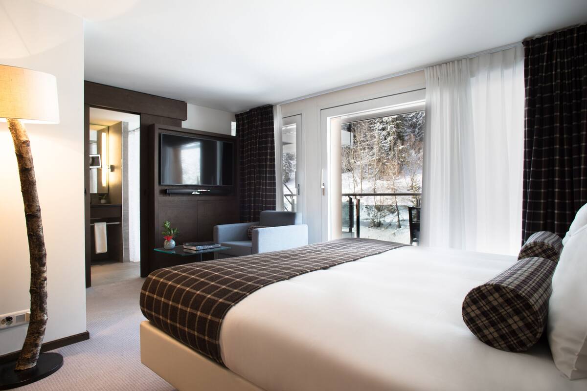 Habitación de hotel moderna con una cama grande, televisión, zona de estar y balcón con vistas a árboles.