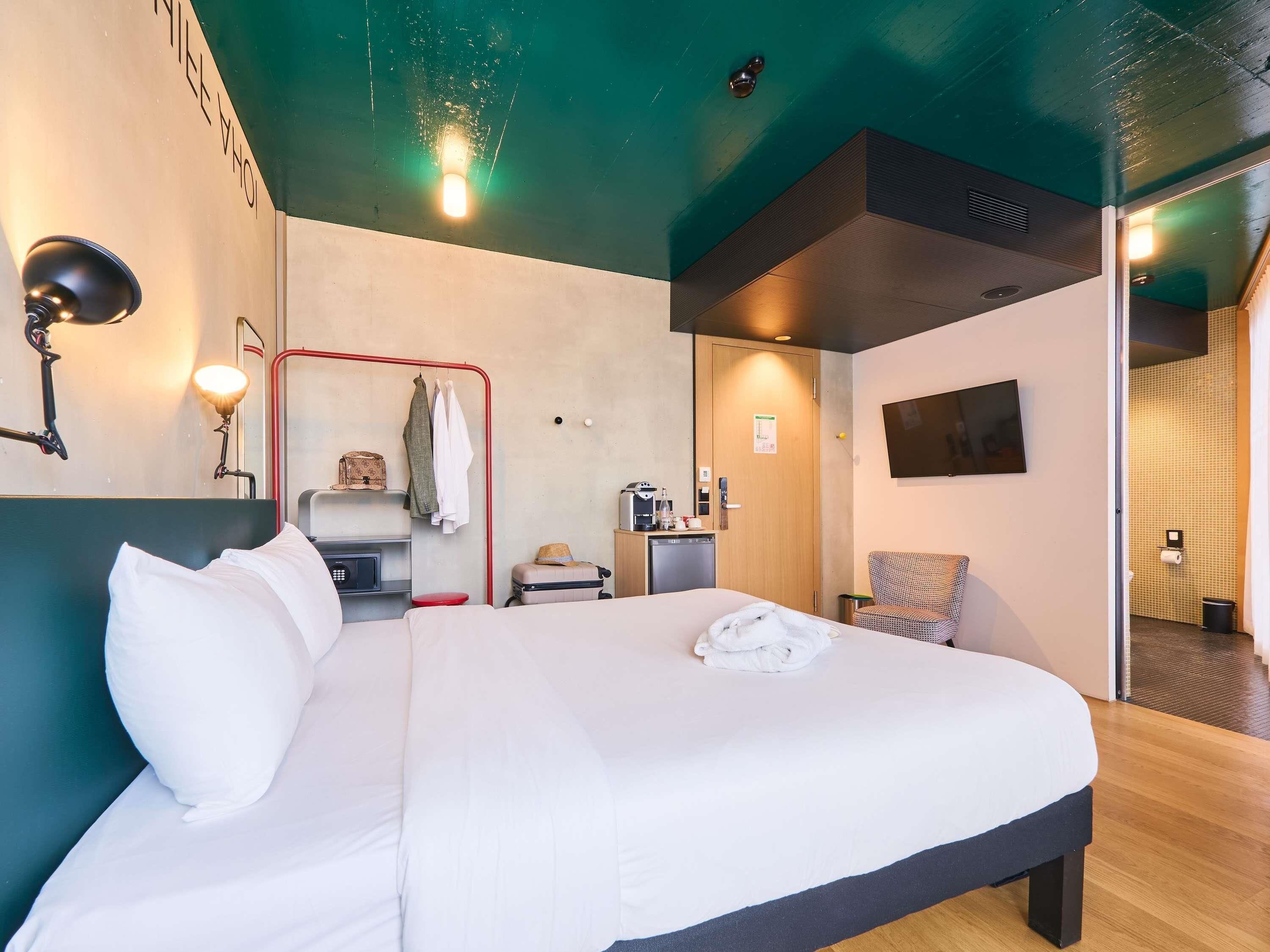 A hotel room with a bed, a desk, a chair, a TV, and a bathroom visible through an open door.