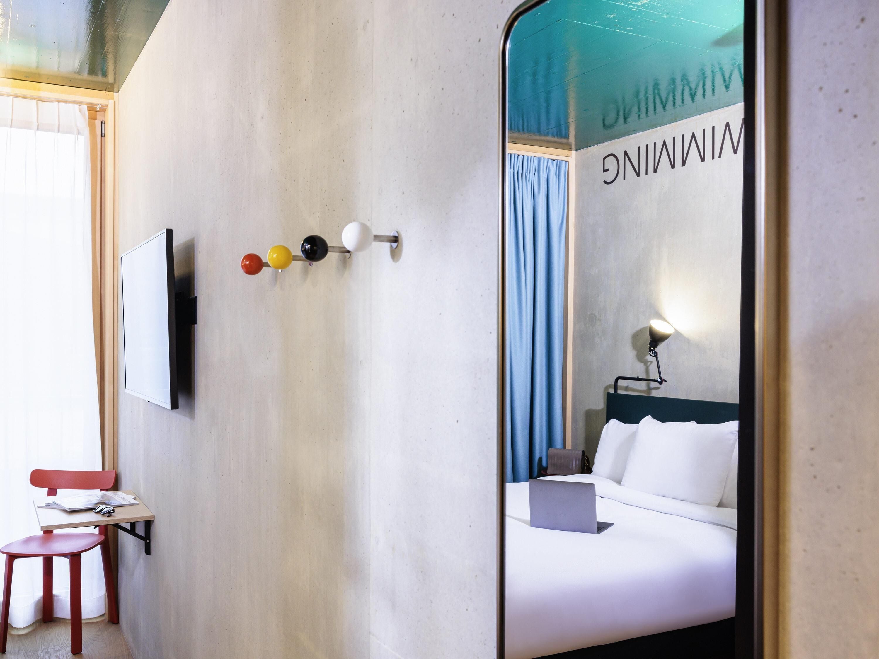 A hotel room with a bed, a desk with a chair, a TV mounted on the wall, and a mirror reflecting the room.