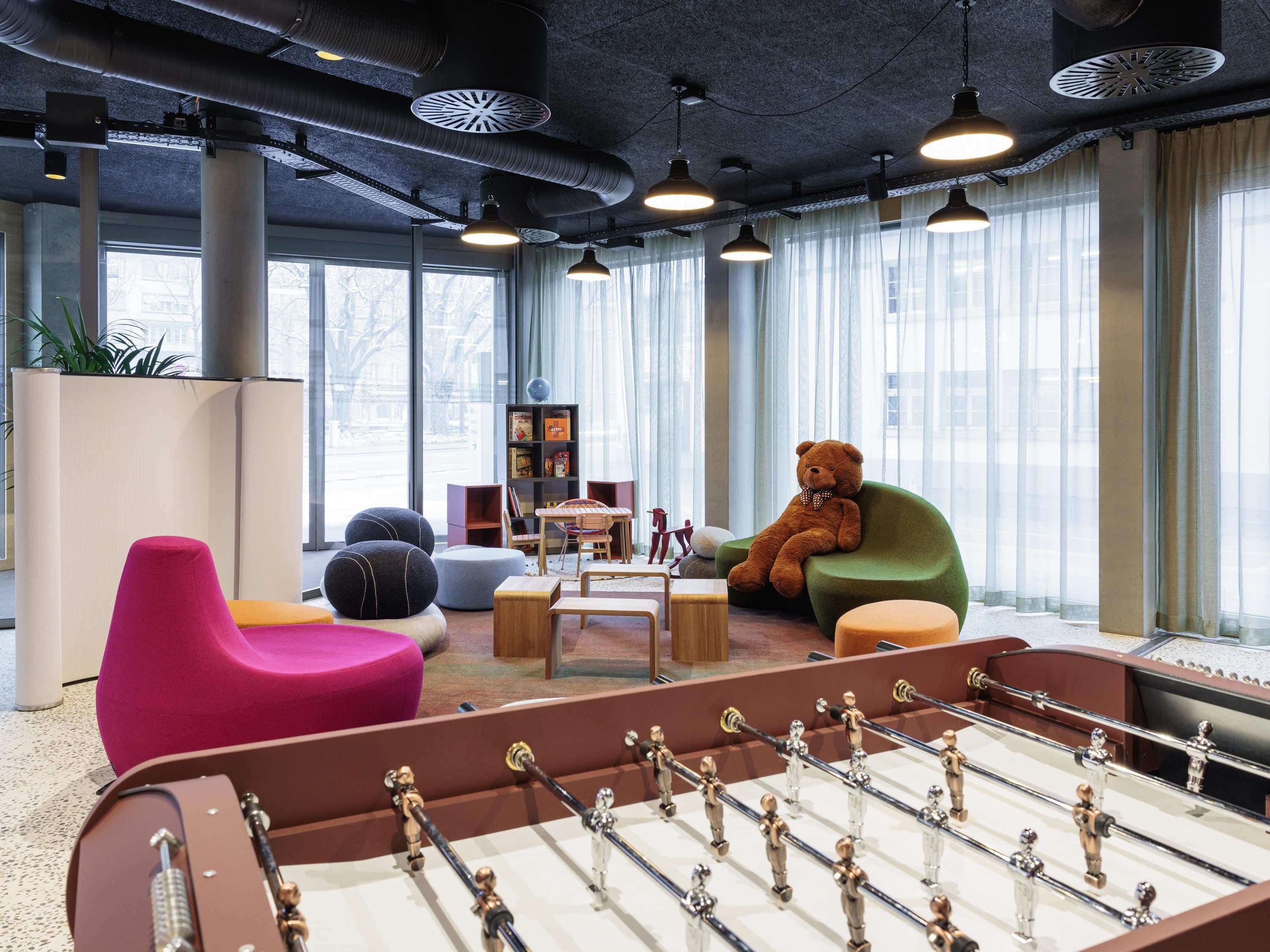 Espaço moderno em ambiente interno com mesa de pebolim, assentos coloridos e grandes janelas.