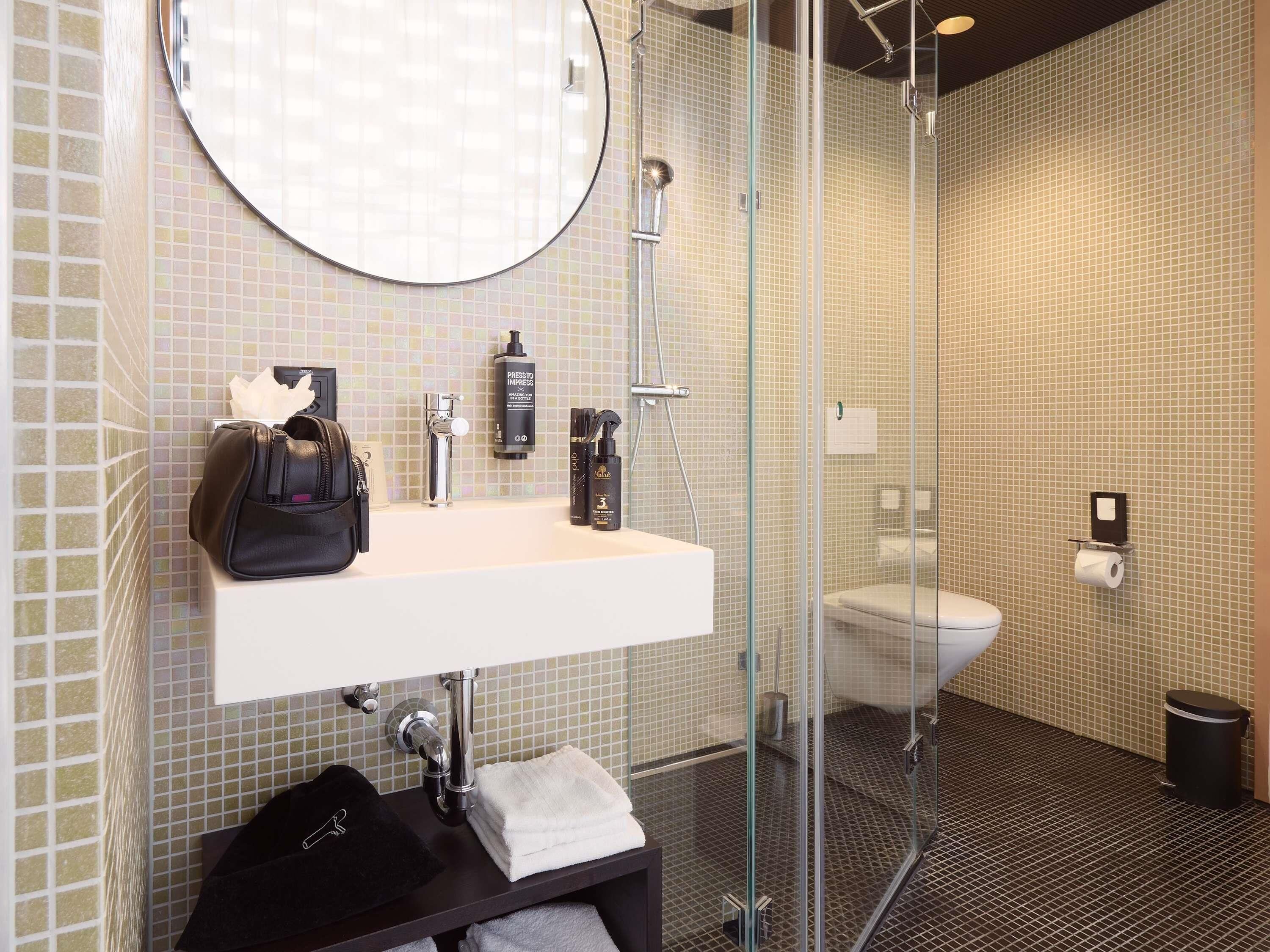 A modern bathroom with a glass-enclosed shower, a round mirror, a white sink, and a black handbag on the counter.